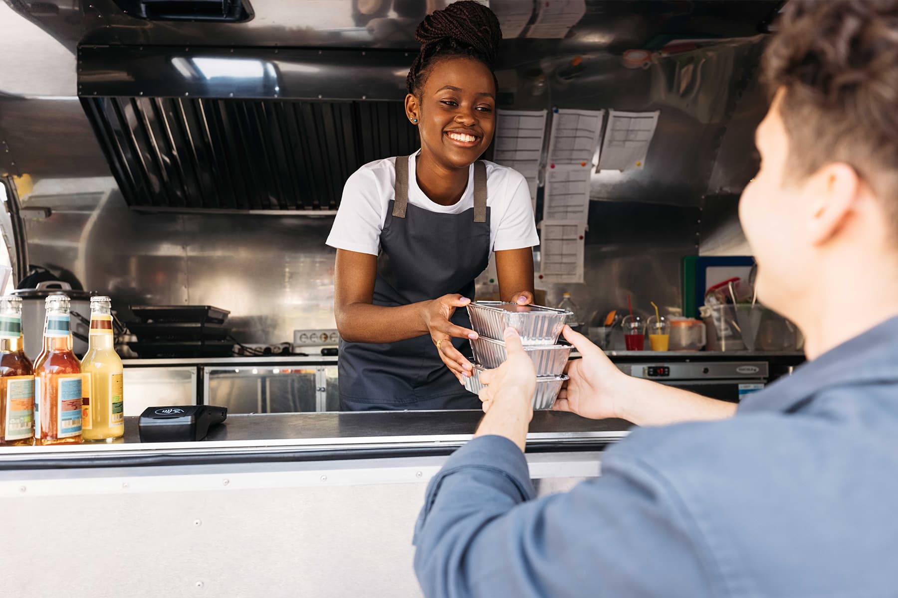 Una mujer con delantal y camiseta blanca entrega una comida en recipientes de aluminio a un cliente desde el interior de un camión de comida. La mujer sonríe afectuosamente. Detrás de ella se ve el interior del camión de comida, junto con salsas variadas y utensilios en el mostrador.