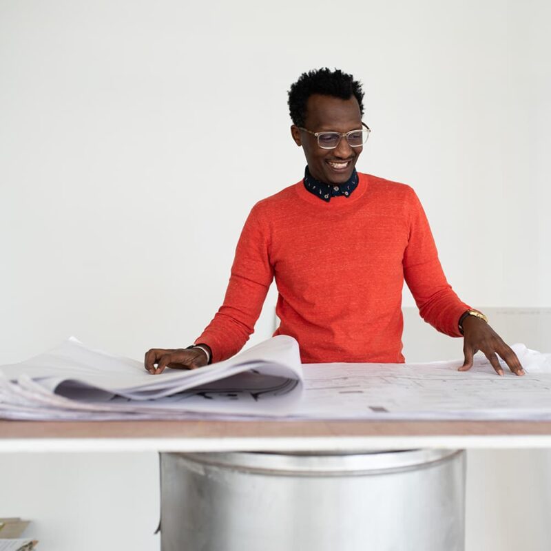 Una persona con gafas y un jersey rojo brillante está de pie ante una gran mesa cubierta de planos arquitectónicos y documentos, sonriendo mientras revisa los papeles. El fondo es liso y blanco, centrando la atención en el área de trabajo del individuo.