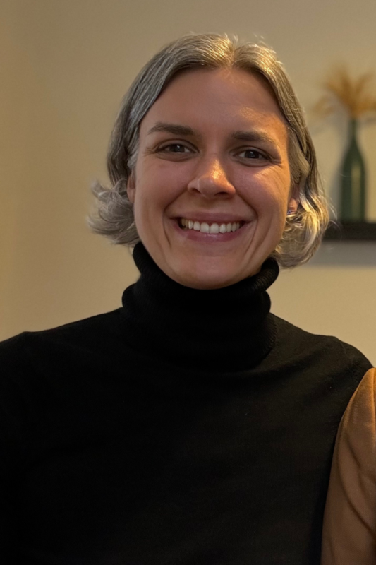 A person with short, gray hair and a big smile is wearing a black turtleneck sweater. They are in an indoor setting with a neutral background, and a decorative vase with dried flowers can be seen in the blurred background.