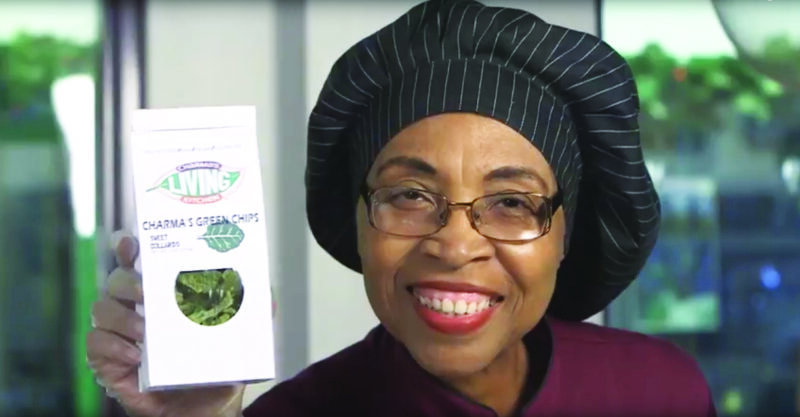 A smiling person wearing glasses and a black chef's hat holds up a white box labeled "Charma's Green Chips." The background shows greenery, suggesting a fresh, healthy setting.