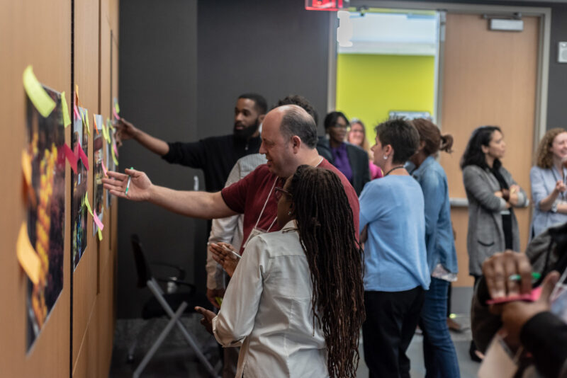 A diverse group of people is gathered in a room, engaged in a collaborative activity. Some are attaching colorful sticky notes to pieces of paper on the wall, while others observe and discuss. The atmosphere appears to be lively and interactive.