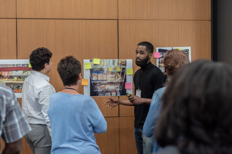 A man stands in front of a group of people, speaking and pointing to a board covered with images and sticky notes. The group is listening attentively, with one person facing the speaker and others slightly turned towards him. They are in a room with wooden panel walls.