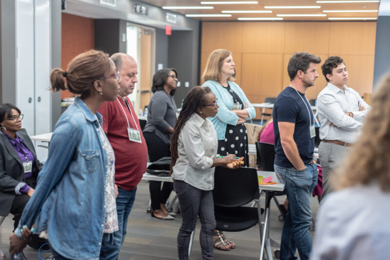 A diverse group of people stands in a classroom or meeting room, attentively facing toward the front. Some are wearing name tags and casual attire, including jeans and shirts. The room has modern decor, with wooden paneling and overhead lights. Tables and chairs are visible.