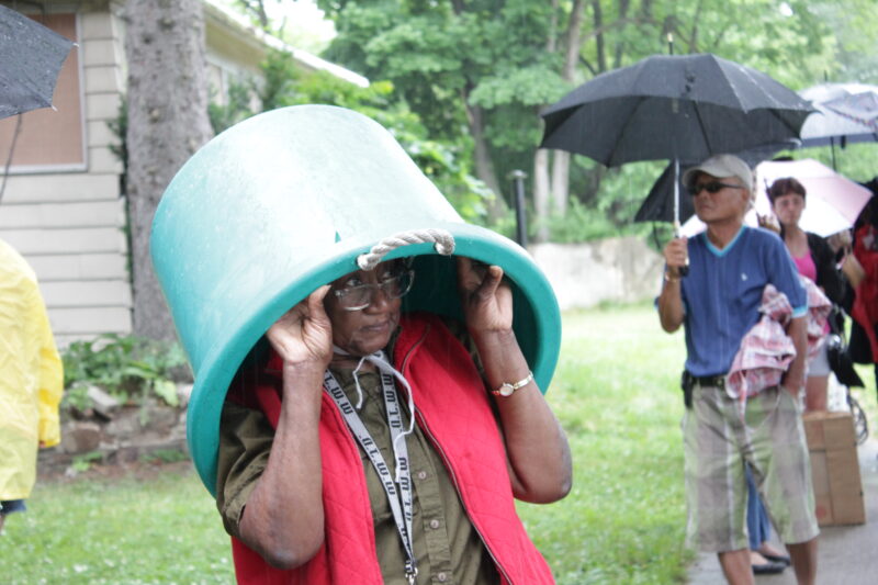 A person wearing a red vest uses a large blue plastic tub as a makeshift hat while walking outside. Several people in the background carry umbrellas, and the scene appears to be in a residential area on a rainy day.