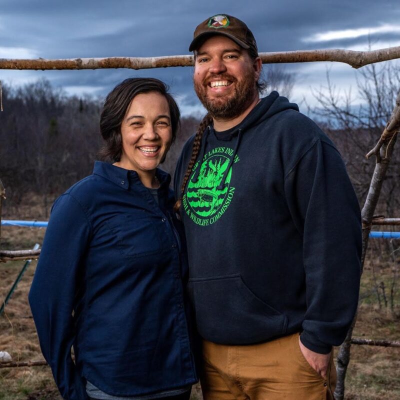 A smiling woman in a blue long-sleeved shirt stands beside a man with a braided beard wearing a green and black cap, a black hoodie, and brown pants. They are outdoors with a wooden structure and a cloudy sky in the background.