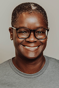 A person with short hair and glasses is smiling at the camera, wearing a gray t-shirt. The background is plain and light-colored.