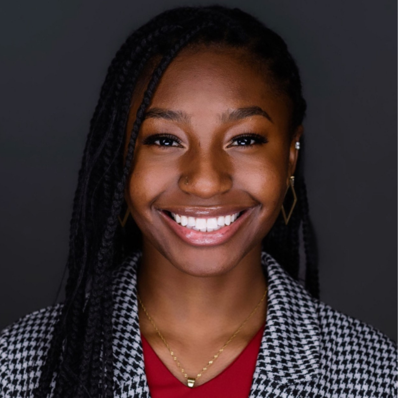 A person with long braided hair, wearing a black-and-white houndstooth blazer over a red top, smiles brightly against a dark background. They are also wearing geometric gold earrings and a gold necklace.