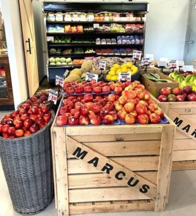 Fruit and vegetable displays are arranged in a grocery store. Baskets and bins are filled with apples, lemons, bananas, and other produce. Shelves in the background showcase packaged goods. A wooden crate labeled "MARCUS" holds apples in the foreground.