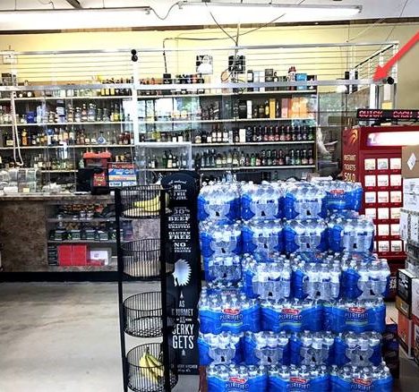 A small convenience store has shelves stocked with various beverages and products. The foreground features cases of bottled water stacked high. To the right, there's a red vending machine. On the left, bananas are displayed in a wire basket.