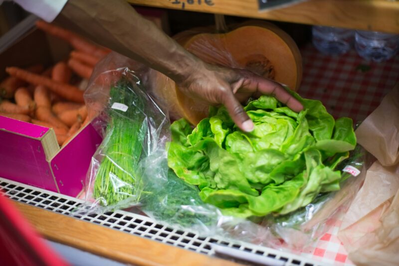 pexels-kampus-8475113 Hand reaching to grab a head of fresh lettuce sitting on a white, metal shelf with other fresh herbs and vegetables surrounding it.