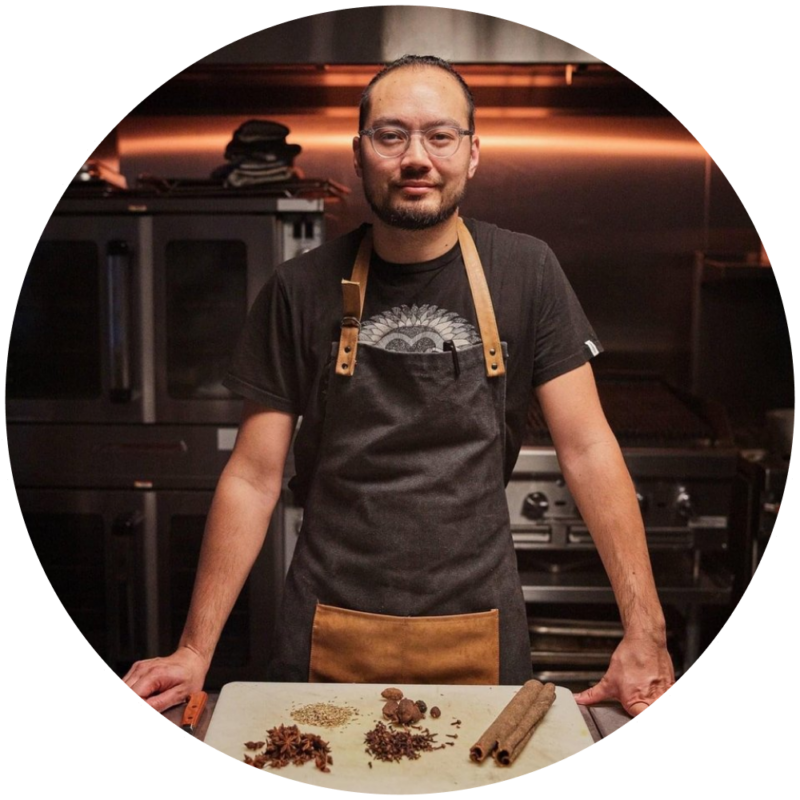 A person wearing glasses, a dark T-shirt, and an apron stands at a kitchen counter with various spices spread out. The kitchen has an industrial design with stainless steel appliances and a warm, dim lighting ambiance.
